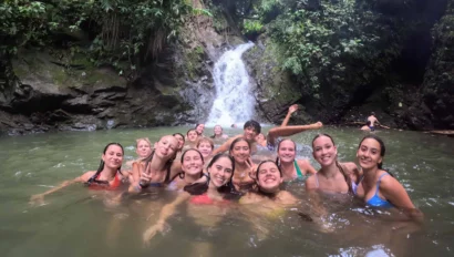 Group of people swimming and posing in front of a waterfall surrounded by lush greenery.