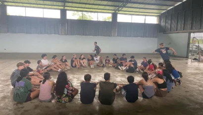 A group of people sitting in a circle on the floor inside a large hall. One person is jumping in the center of the circle.