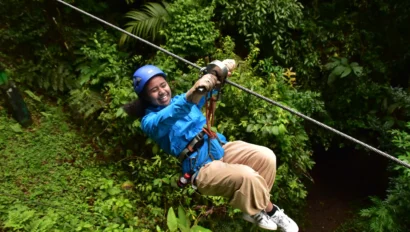 Person ziplining through lush green forest, wearing a blue helmet and jacket, with a joyful expression.