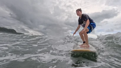 A teen enjoys a summer trip, riding a wave on a surfboard under a cloudy sky, with the ocean and distant shoreline visible.