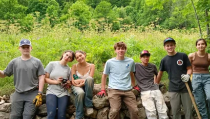 Seven young people in outdoor work clothes sit and stand by a low stone wall in a green, wooded area, with tools and gloves visible—capturing the spirit of a Costa Rica student travel adventure.