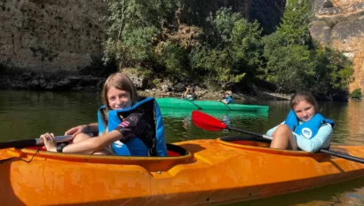 Two children in blue life jackets paddle an orange kayak on a calm river, surrounded by rocky cliffs and trees.