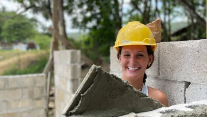 A student traveler wearing a yellow construction helmet smiles while applying mortar to a concrete block wall at a construction site, possibly as part of a school group travel experience. Trees and unfinished walls are visible in the background.