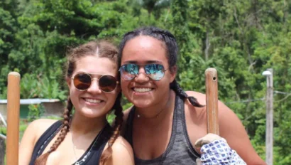 Two student travelers wearing sunglasses and gloves stand outdoors holding wooden poles, smiling at the camera with a green, forested background—enjoying a refreshing break during their school group travel adventure.