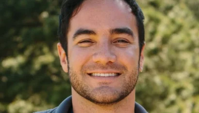 A man with dark hair and a beard smiles at the camera, standing outdoors with trees in the background.