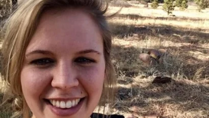 A person smiling in a grassy field with trees and mountains in the background.