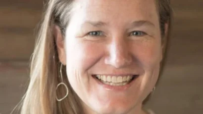 Smiling person with long hair and hoop earrings against a wooden background.