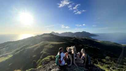 Four people sit together on a rocky hilltop, overlooking a scenic landscape with the sun shining brightly in the sky.
