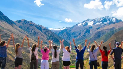 A school group travel program by Global Works brings together adventurers standing on a mountain trail, raising their arms toward snow-capped peaks and a sweeping valley beneath a clear blue sky.