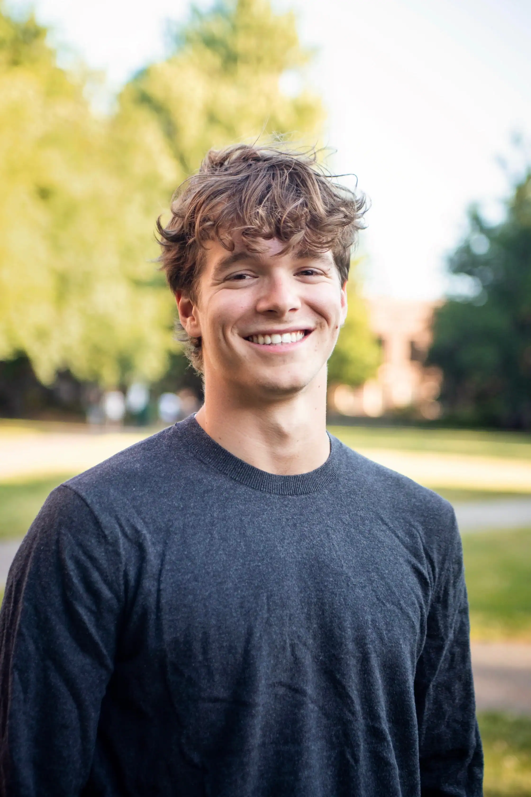 A young man with wavy brown hair, wearing a dark gray sweater, stands outside smiling, with green trees and a blurred building in the background.