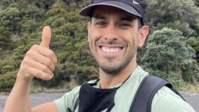 A person outdoors, smiling and giving a thumbs-up, wearing a black visor and backpack, with a green forest background.