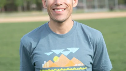 A person standing outdoors on grass, wearing a blue t-shirt with a colorful graphic and the text "GLOBAL WORKS TRAVEL WITH PURPOSE." Trees and mountains are visible in the background.