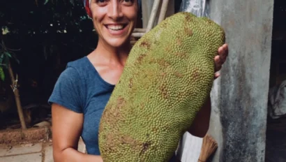 A person smiling and holding a large, green jackfruit with a textured surface, standing outdoors.