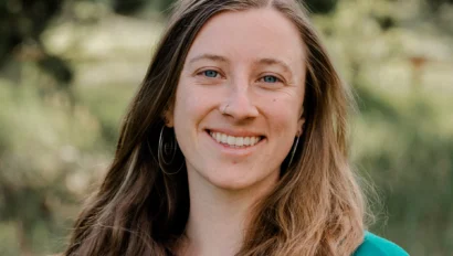 Woman with long hair wearing a turquoise top stands outdoors with greenery in the background.