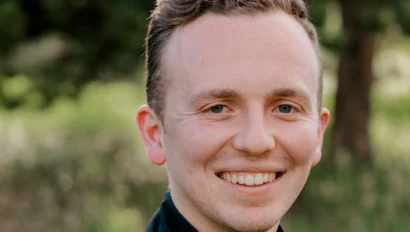 A person in a black jacket smiles outdoors with greenery in the background.