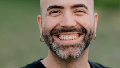 A man with a beard and hoop earrings smiles broadly. He is wearing a black Volcom Stone T-shirt. A blurred green background is visible.