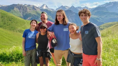 A group of five people smile and pose together on a grassy hillside, with a mountain range in the background under a clear blue sky.