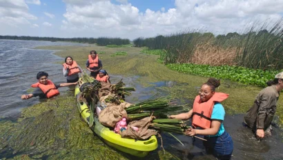 Five students in life jackets, possibly on USA school trips, gather aquatic plants and load them into a kayak while standing in shallow, vegetation-filled water under a partly cloudy sky.