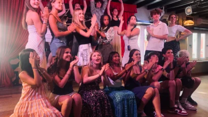 A group of young people pose and clap together on a stage, some standing and some sitting, with red curtains and spotlights in the background—capturing the excitement of their Spain school trip.