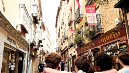 A group of young people with backpacks walk down a narrow street lined with shops and balconies during a school trip in Spain.