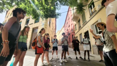 A group of students stand in a circle on a sunny street lined with colorful buildings, listening to a guide under the shade of green trees during their Spain school trip.