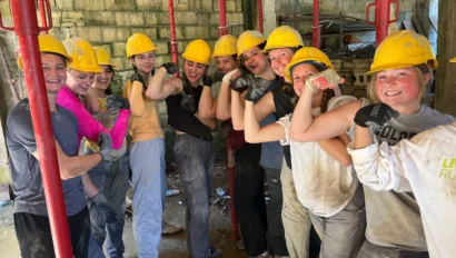 A group of young student travelers in yellow hard hats and work clothes pose together indoors, flexing their arms and smiling during a Puerto Rico service trip in a construction setting.