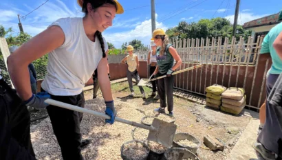 Several people on a Puerto Rico student service trip, wearing helmets and gloves, are shoveling gravel into buckets outdoors on a sunny day, next to a fence and bags of materials.