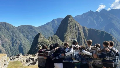 A group of students stand arm in arm facing Machu Picchu, with green mountains and a clear blue sky in the background, capturing the spirit of a memorable Peru school trip.