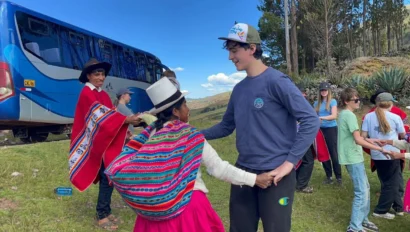 A group of people dance outdoors near a parked blue bus during a Peru school trip; one wears traditional Andean clothing, while others dress casually.