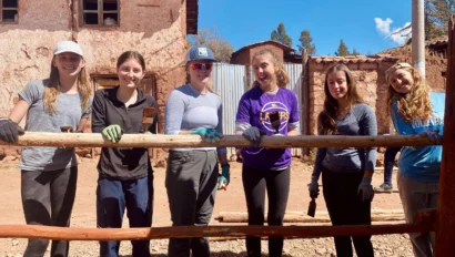 Six students stand outdoors in front of a rustic building during their Peru school trip, smiling and holding onto a wooden fence, wearing casual clothes and work gloves.