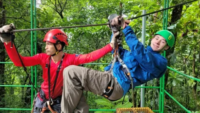 Two people in helmets and harnesses ziplining through a lush forest, with one person guiding the other on the platform.