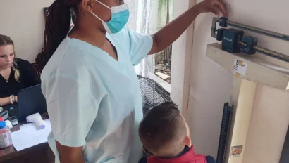 A nurse measures a young boy's height using a wall-mounted scale. The boy faces away from the scale while the nurse adjusts the measurement bar. A person sits nearby at a desk.