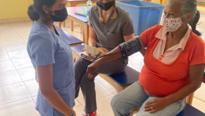 Two healthcare workers wearing masks check the blood pressure of an older woman in a bright room. One kneels while holding the cuff, and the other observes.