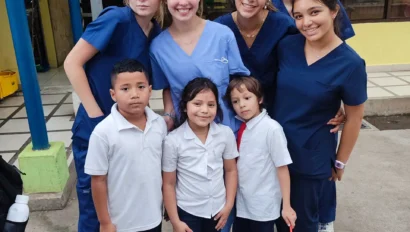 Group of students in scrubs posing with three children in school uniforms outside a building.