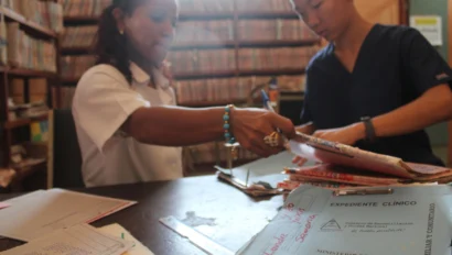 Two people organize folders and documents at a table in a room filled with shelves of files.