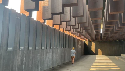 A person walks alone through an outdoor memorial with suspended metal columns and engraved plaques on the walls, exploring a powerful site of American history travel under natural daylight.