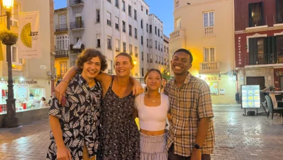 Four young adults stand together smiling in an outdoor plaza at dusk, enjoying their language immersion trip with buildings and streetlights glowing in the background.