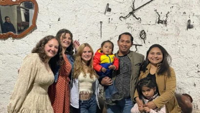 Six people, including two small children, pose for a photo during their language immersion trip, standing against a textured white wall decorated with rustic tools and a mirror.