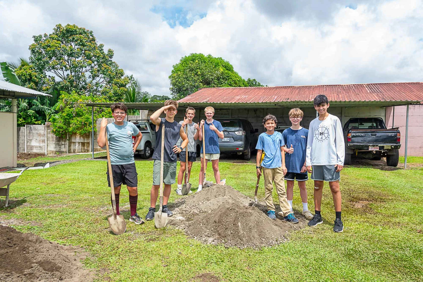 A group of seven people stand on grass holding shovels near a pile of dirt. Cars and trees are in the background.