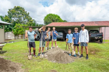 A group of seven people stand on grass holding shovels near a pile of dirt. Cars and trees are in the background.