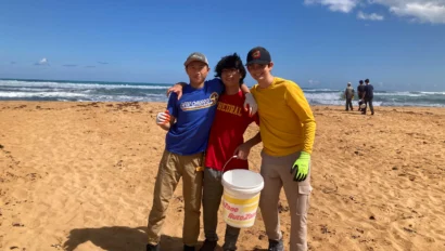 Three people stand on a sandy beach, arm in arm, holding a bucket. The ocean is visible in the background under a blue sky with clouds.