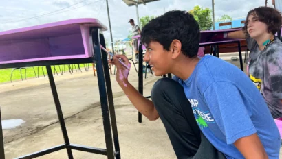 A boy in a blue shirt paints the leg of a pink desk outside under a metal roof during one of the Middle School service trips, while another person sits nearby.