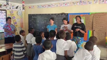 A group of children in school uniforms gathers around three students at the front of a classroom with a chalkboard and colorful decorations during a teen program that works with kids.