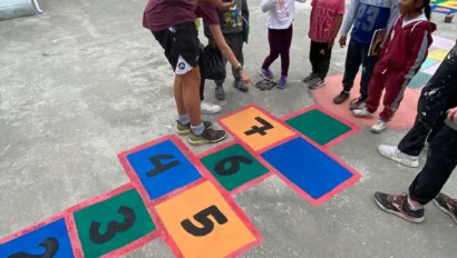 A group of children and an adult stand around a colorful hopscotch grid painted on a concrete surface in an outdoor area, similar to activities offered by teen programs that work with kids.