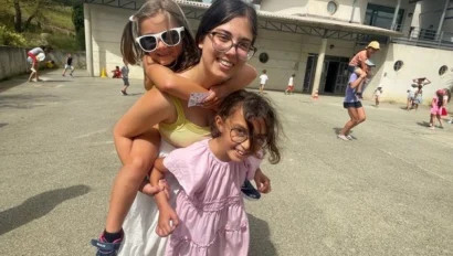 A young woman, part of teen programs that work with kids, carries a smiling child on her back while standing next to another young child in a pink dress outdoors near a building with others in the background.