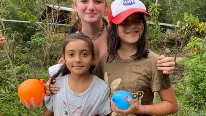 Three girls stand outdoors holding water balloons, with greenery and trees in the background. Two are wet and smiling, while one stands behind them with her arms around their shoulders—capturing the fun of teen programs that work with kids.