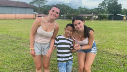 Two women and a boy smiling and posing together on a grassy field with a building and trees in the background.