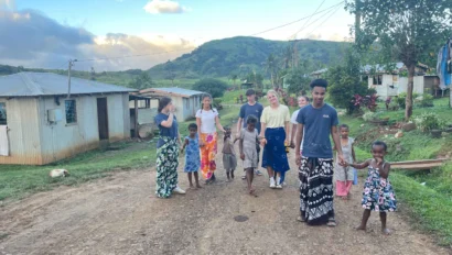 A group of people, including children, walk along a dirt road in a rural area with houses and green hills in the background.