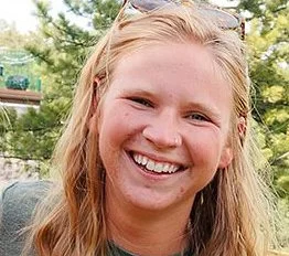 A person with long blonde hair smiles outdoors, with trees in the background.
