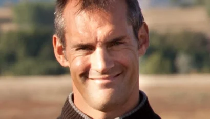 A man in a brown zip-up sweater smiles in an outdoor setting with trees and hills in the background.